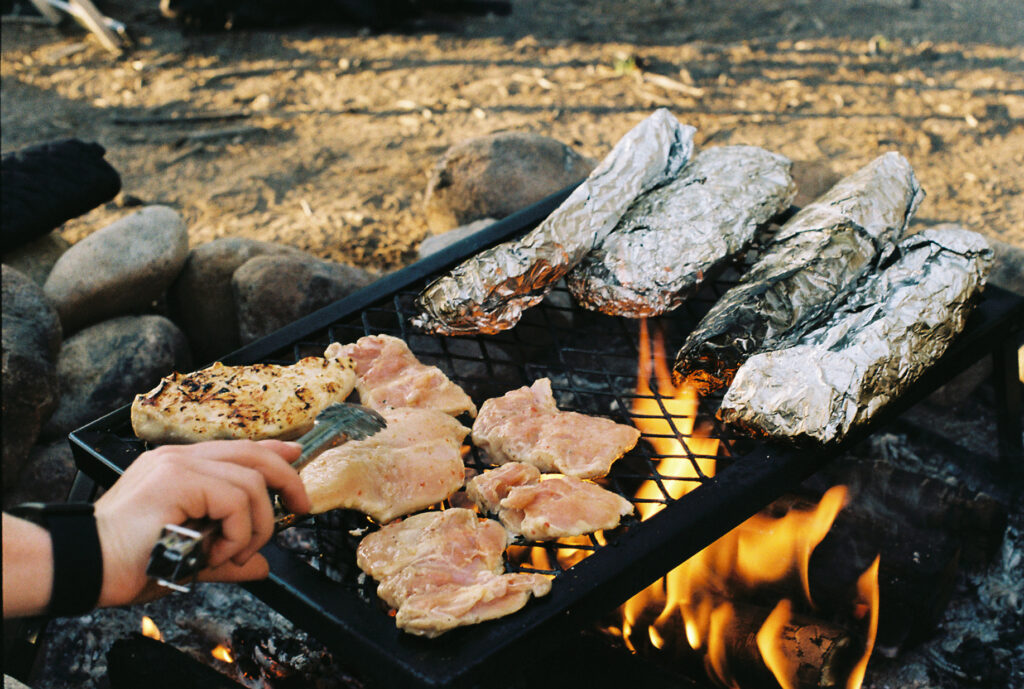 A grill over a campfire cooking chicken and veggies in tin foil.